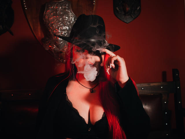 AprilDoel with red hair, black hat, and smoking, wearing a black top with deep cleavage, holding her hat, showing her tits, in a dim red-lit room.