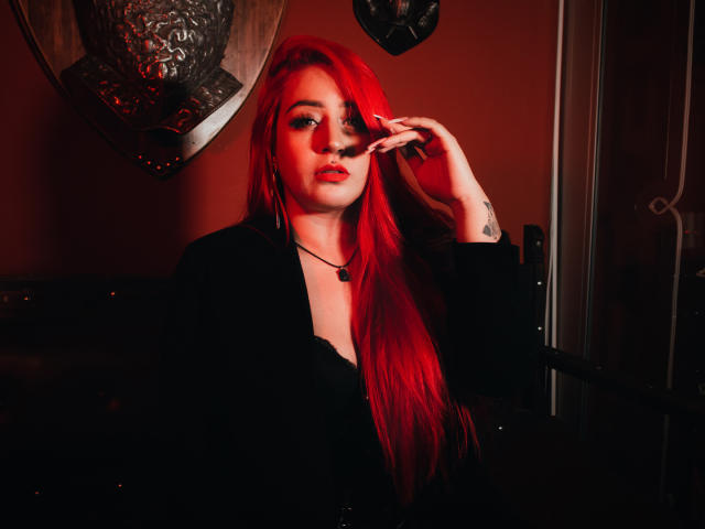 AprilDoel, con lunghi capelli rossi, giacca nera e collana nera, che si tocca il viso, illuminazione rossa, tatuaggio sul polso destro, che guarda la fotocamera, sexy, attraente..