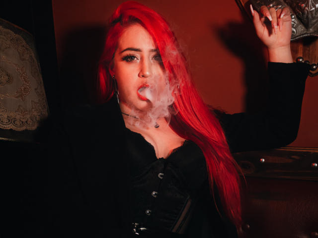 April Doel, con el cabello rojo brillante, fumando un cigarrillo, vestida con un corpiño negro, con los senos prominentes, sosteniendo un vaso en su mano derecha, con lápiz labial rojo y ojos delineados..