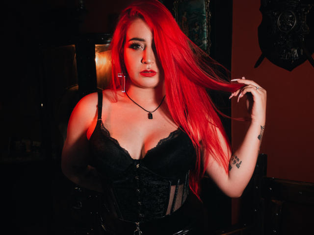 AprilDoel, con i capelli rosso acceso, un corpetto di pizzo nero e una collana nera, si tiene i capelli, mostrando tatuaggi sul braccio destro, con un'illuminazione rossa sul viso..