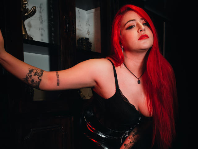 AprilDoel, avec ses cheveux roux, son haut en dentelle noire et son pantalon en cuir noir, appuyée contre du bois sombre. Elle a un tatouage sur le bras droit, du rouge à lèvres et un collier noir. Ses seins sont visibles..