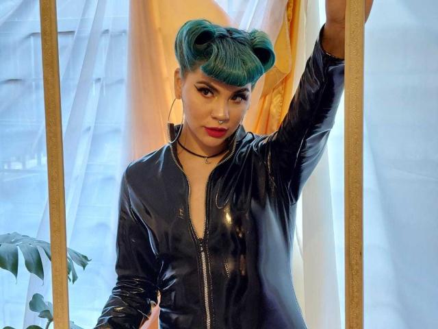 AndiChin with teal pigtails, red lipstick, black shiny leather zip-up, arm raised, standing in front of white and orange curtains, wooden frame on sides.