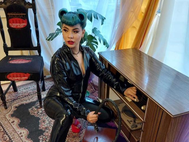 AndiChin, avec ses cheveux turquoise, son rouge à lèvres rouge et sa tenue en latex noir, est agenouillée près d'un meuble en bois, tenant des bottes en cuir noir, et montrant son pantalon en latex noir brillant, ainsi que ses petites seins noirs..
