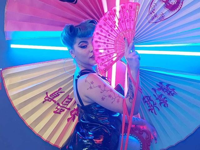 AndiChin with blue hair, holding pink fan, wearing shiny black dress, tattoos on arm, looking back, neon blue and pink lights, big hoop earrings, fan has Chinese characters.
