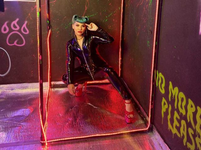 AndiChin, vêtue d'une combinaison en latex noir brillante, est accroupie dans une boîte éclairée au néon, portant des talons rouges, avec des cheveux bleu-vert, une main sur la tête, et une expression séduisante, mettant en valeur ses fesses..