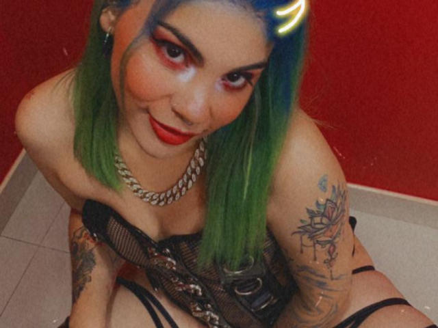AndiChin with green hair, red lipstick, and a neon horn headpiece, wearing a black mesh corset and chain necklace, has tattoos and is kneeling with her tits visible.