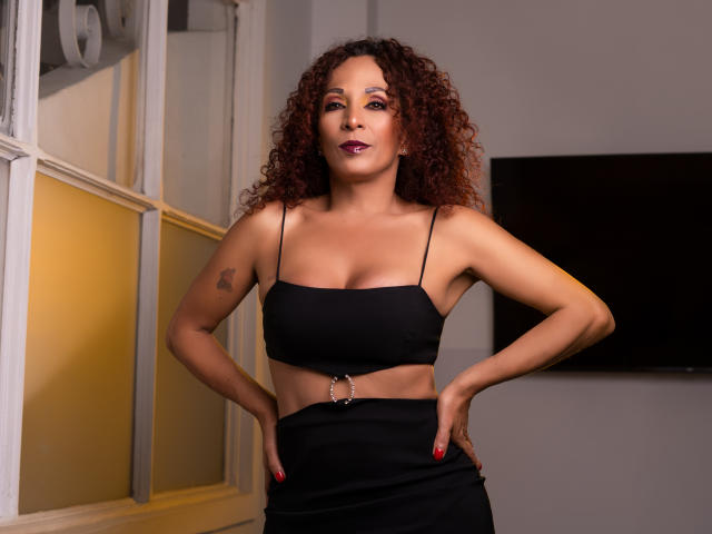 KhazandraX with curly red hair, tan skin, small tattoos, black spaghetti strap crop top, black skirt, red nails, hands on hips, confident smile, medium breasts, metal belt ring.
