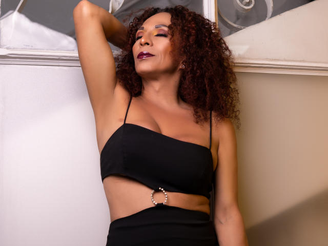 KhazandraX with curly red hair, wearing a black crop top, showing her tits, with a ring on her midriff, arm raised, eyes closed, and dark lipstick.