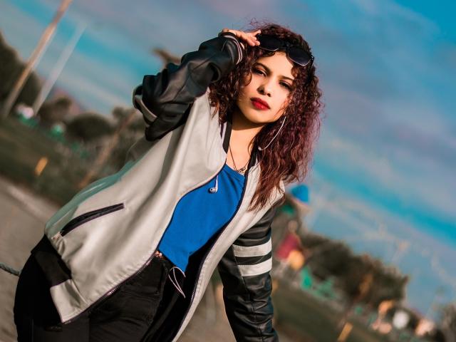 Ariana, atractiva, con cabello castaño rizado, vistiendo una chaqueta gris y negra, una camisa azul, pantalones negros y gafas de sol en la cabeza, de pie al aire libre, con la mano en el cabello, mirando a la cámara, labios rojos..