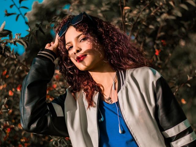 Ariana, uma mulher atraente com cabelos cacheados, vestindo uma jaqueta preta e branca, uma camisa azul e batom vermelho, segurando óculos de sol contra um fundo ensolarado e cheio de folhagens. Ela está sorrindo e usa um piercing no nariz..