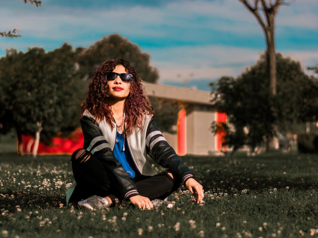 Die lockenköpfige Schönheit Ariana sitzt auf dem Gras, trägt eine Sonnenbrille, ein blaues Hemd, eine schwarze Jacke und eine schwarze Hose, lächelt entspannt und sitzt mit verschränkten Beinen in einem sonnigen Park..