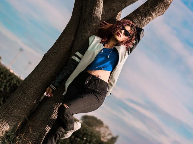 Hottie Ariana lehnt an einem Baum, trägt ein blaues Crop-Top, schwarze zerrissene Jeans und eine weiße Jacke, hat lockiges rotes Haar und eine Sonnenbrille und sieht sexy und entspannt aus..