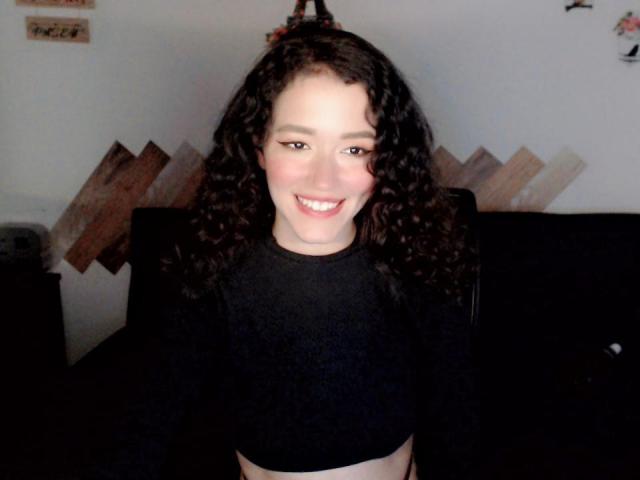 RukkiaParist met krullend donker haar, lachend, draagt een zwarte crop top, waarbij haar borsten zichtbaar zijn, en staat voor een zwarte leren stoel..