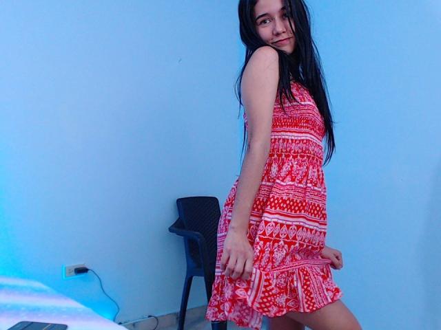 DeisyWest with long black hair, red and white patterned dress, standing sideways, showing her ass, looking over shoulder, in a blue-walled room, black chair behind her.