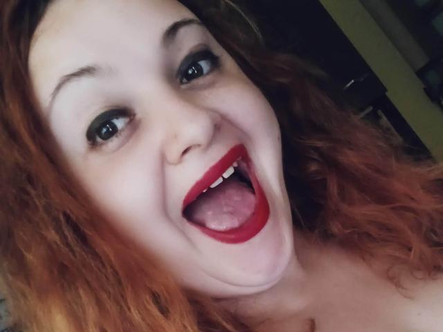 O rosto de CurlyCherry, com a boca bem aberta e batom vermelho, a língua visível, olhos azuis, cabelo laranja cacheado, sorrindo, pele clara, ligeiramente cheinha, nariz com piercing, sobrancelhas escuras, com uma expressão feliz..