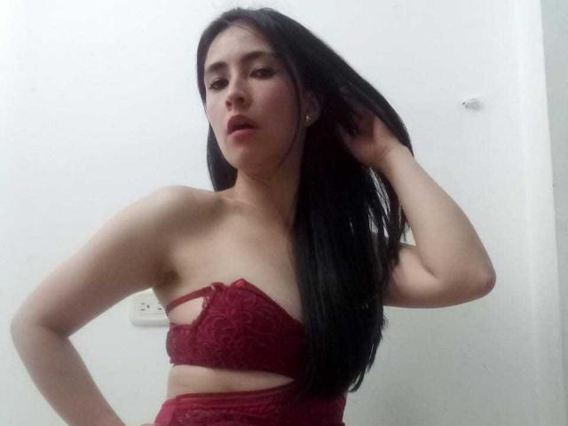 JainaRogers with long black hair, wearing a red lace bra that shows her tits and a matching red lace bottom, standing with one hand in her hair.