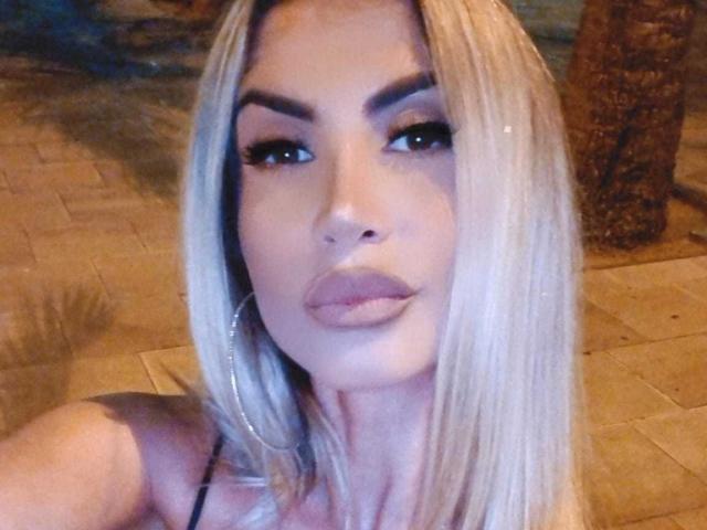 Lellie, con i capelli biondo platino, sopracciglia folte e labbra piene di colore naturale, indossa grandi orecchini a cerchio e guarda la fotocamera con un sorriso sottile e sicuro..