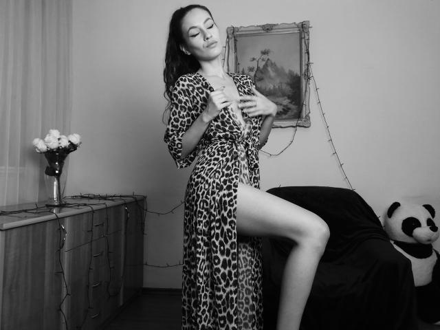 VievienneClemente stands in a black-and-white photo, wearing a leopard print dress, hands on chest, leg raised, showing her pussy. She has closed eyes and a sultry expression.