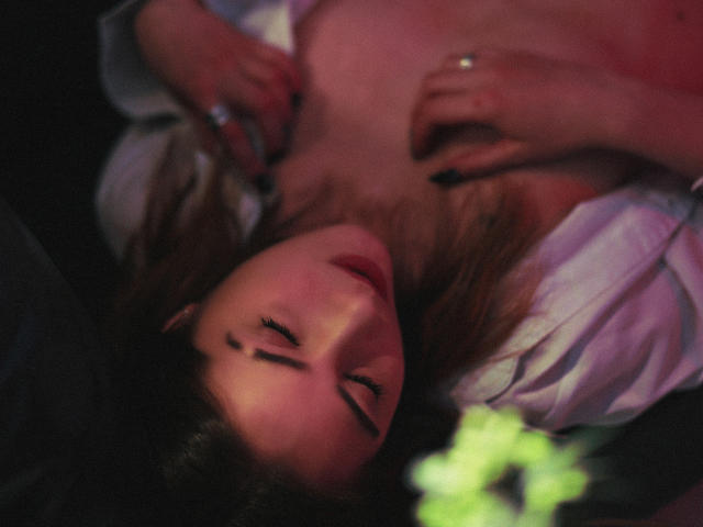 Decadance's face, eyes closed, lying down with brown hair. Her hands rest on her chest, exposing her small tits. Soft pink and red lighting. Gentle, intimate, sexy.