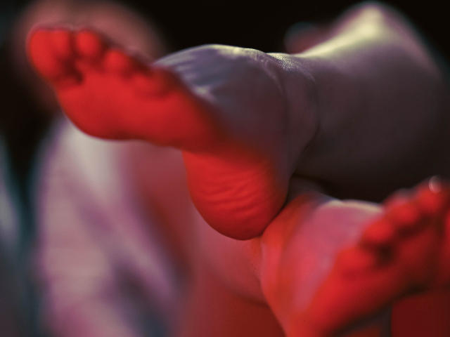 Decadance's feet, red-soled with visible toe wrinkles, are prominently in the foreground, slightly blurred, with a soft, pinkish glow on the soles and toes.