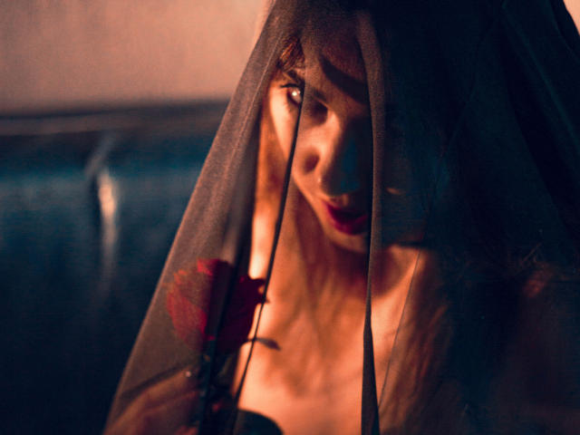 Decadance's face is partially covered by a sheer black veil, with a red rose in the foreground, her expression is intense and her bare shoulders are visible.