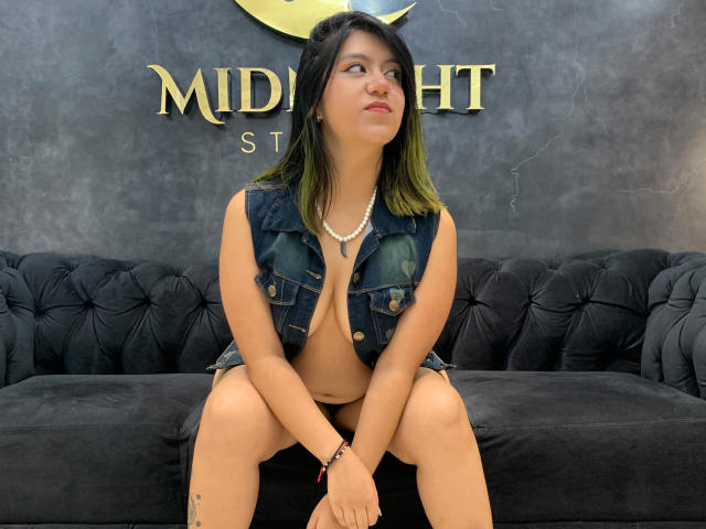 MonserrathMoon with green-tipped black hair, wearing a sleeveless denim vest and pearl necklace, sitting on a black tufted sofa, exposing her bare tits and pussy.