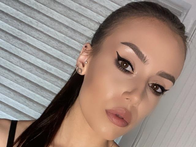 VannaBlack hat braune Augen, einen dicken schwarzen Eyeliner, volle, braue Augenbrauen, einen dezenten rosafarbenen Lippenstift und einen glatt zurückgebundenen Pferdeschwanz mit kleinen Ohrsteckern. Ihr Gesicht ist leicht nach rechts geneigt..