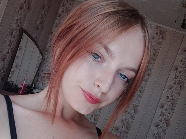 Markizza, con capelli rossastri, occhi azzurri e rossetto rosso, sorride leggermente, indossando un reggiseno nero a spalline, in un selfie. Il suo seno piccolo è appena visibile..