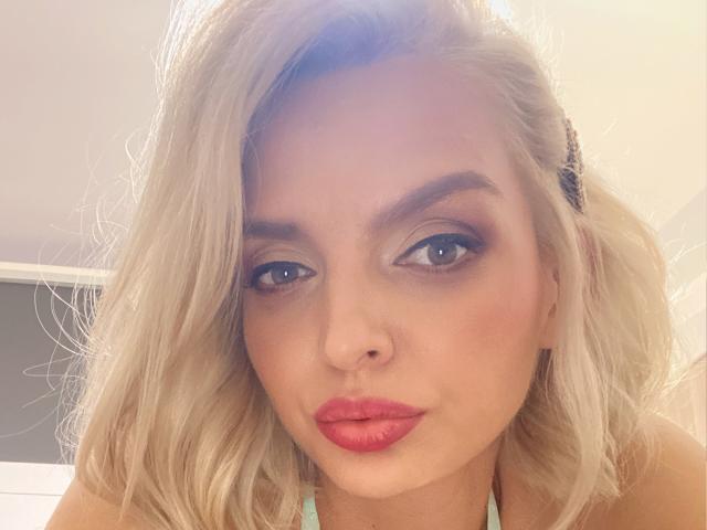 DouceAmelia has blonde hair, blue eyes, and red lipstick. She's making a pouty face with her nose slightly up. Her tits are not visible in this shot.