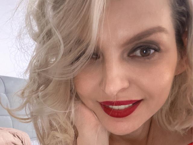 DouceAmelia has blonde curly hair, red lipstick, and a cute smile. Her brown eyes are slightly squinted. She's wearing a pink shirt. Her tits are not visible.