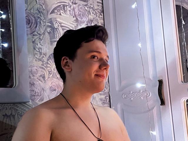 Shirtless LonelyWolf with short dark hair, necklace, smiling at camera, white door with string lights, patterned wallpaper, soft lighting, slightly chubby face, visible nose, relaxed expression.
