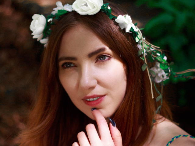 Decadance has pale skin, green eyes, and brown hair, wearing a white rose and green leaf headband, with her finger touching her pink lips, showing dark nail polish.