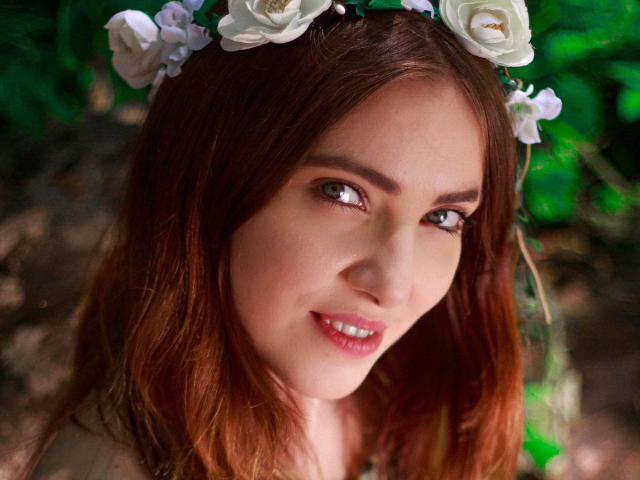 Decadance has light brown hair, a white flower crown, and a cute smile with pink lips. Her brown eyes look slightly to the side, showing a pretty face.