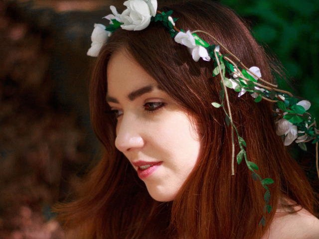 Decadance ha la pelle chiara, i capelli castani e una corona di fiori bianchi con foglie verdi. Guarda di lato con un sorriso dolce, mostrando le labbra rosate..