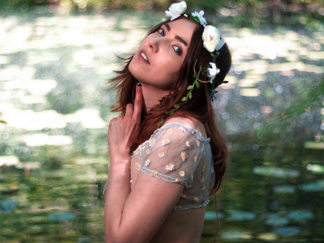 Decadance with medium brown hair, white flower headpiece, and sheer top with daisy patterns, touching her neck, looking back with a slight smile, near a reflective water surface.