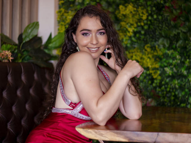 NalimiSmirnov, avec ses cheveux bruns bouclés, souriante, portant une robe rouge avec des détails argentés, tenant un téléphone, assise dans une cabine en cuir marron, montrant ses fesses..