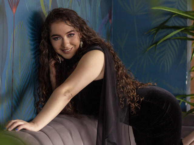 NalimiSmirnov, con i capelli castano ricci, sorridente, indossa un vestito nero, è appoggiata a un divano, mostra il sedere, in una stanza con motivi blu e una pianta verde..