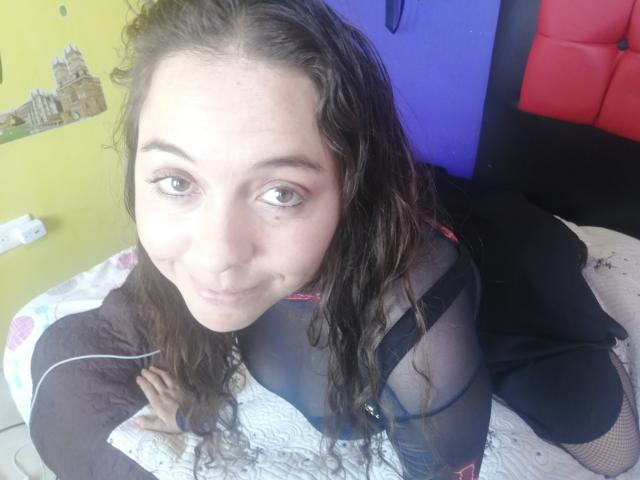 NalimiSmirnov, con i capelli castano ricci, indossa un abito nero trasparente, guarda verso l'alto la fotocamera, è seduta su un letto con lenzuola bianche, sorride, mostrando piccoli seni..