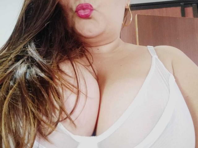 HotValeryPassione avec du rouge à lèvres rose, faisant la moue, portant un soutien-gorge en dentelle blanche, montrant ses grandes seins, de longs cheveux bruns et des boucles d'oreilles dorées, dans un autoportrait légèrement incliné..
