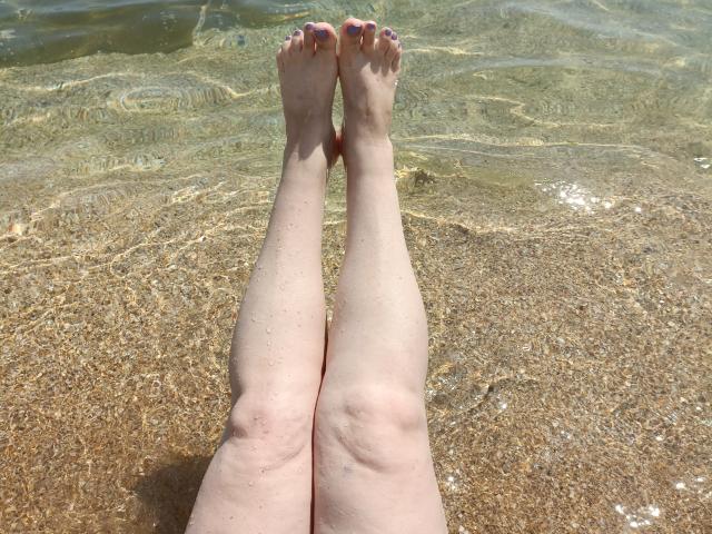 Las piernas pálidas y las uñas de los pies rosadas de StellaDivafox se extienden en agua clara y poco profunda, con los pies ligeramente separados, mostrando suaves ondulaciones y un fondo arenoso..