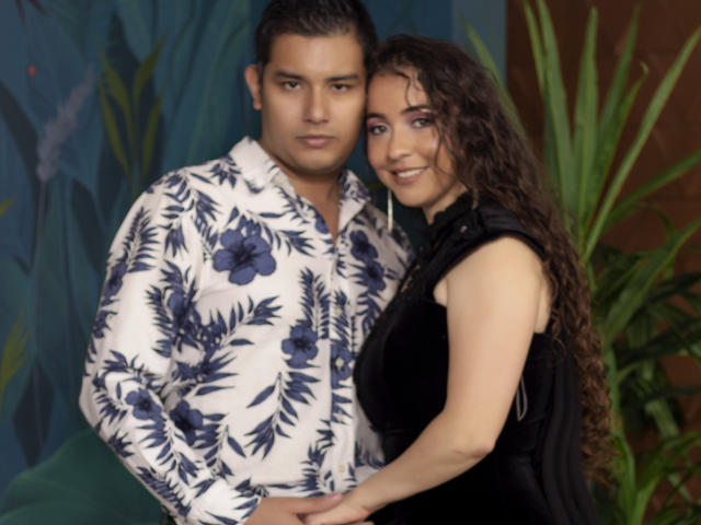 Lucy et Jean, avec des cheveux bruns bouclés, Lucy portant une robe noire sans manches, se tiennent à côté de Jean, qui a les cheveux courts et foncés et porte une chemise à motifs floraux. Toutes les deux sourient à l'appareil photo..