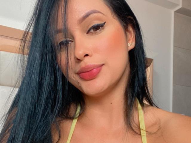 EmilyParkerr ha lunghi capelli neri, eyeliner, rossetto rosa e un sorriso discreto. Indossa un top giallo a halter che mette in mostra il suo seno. È carina e ha un aspetto sensuale..