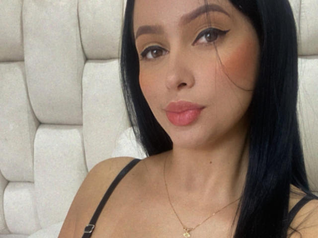 EmilyParkerr with long black hair, brown eyes, and pink lips, wearing a black strap top, and a gold necklace, showing off her small tits and subtle makeup.