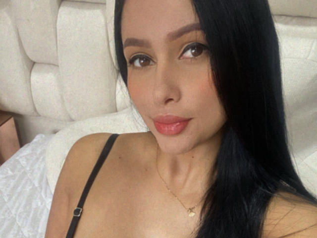 EmilyParkerr with long black hair, brown eyes, and pink lips, wearing a black strap top, lying on a white bed, small gold necklace, subtle makeup, sexy.