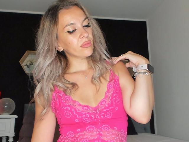 SophieDeee with blonde hair, pink lace top, and winged eyeliner, touching her hair with a pink smartwatch, showing her tits and a subtle necklace.