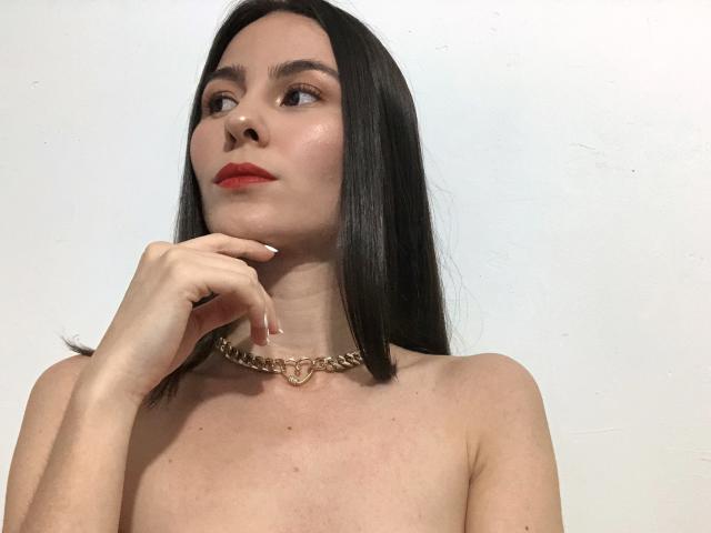 AlisonMeyer with long black hair, red lipstick, and a gold chain necklace, touching her chin, topless, showing her bare shoulders and a slight glow on her skin.