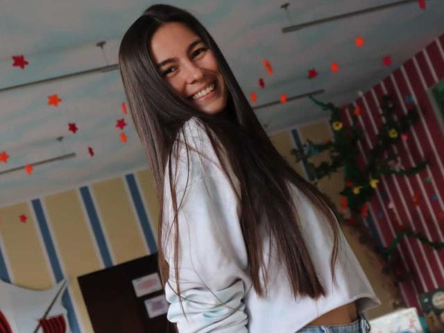 Alison Meyer, com cabelos castanhos longos, sorrindo, vestindo uma cropped branca, olhando por cima do ombro, mostrando o bumbum, em um quarto com decorações coloridas..