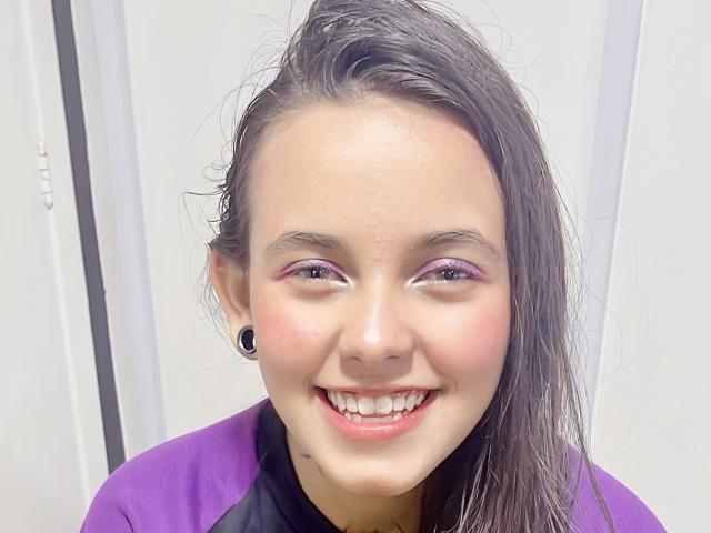 Cloe Ferrara ha i capelli castano scuro e umidi, ombretto viola, espansori per le orecchie neri e una maglietta viola. Sta sorridendo, mostrando i denti, e ha una piccola macchia di acne sul mento..