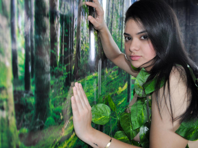 DulcesMiel with long black hair, green leaf in her mouth, hand against forest wallpaper, other hand raised, looking at camera, subtle makeup, gold bracelet, forest background.
