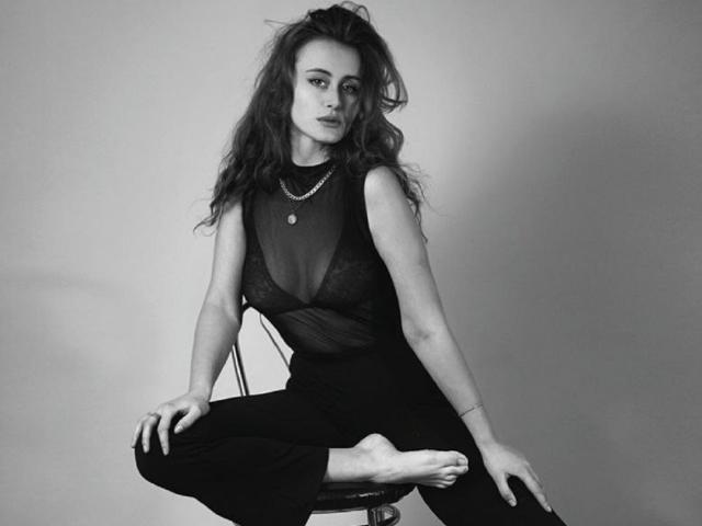 Alesya Drons está sentada en un taburete, vistiendo una blusa negra transparente que deja ver sus senos, pantalones negros y un collar, con un pie levantado, en una fotografía en blanco y negro..